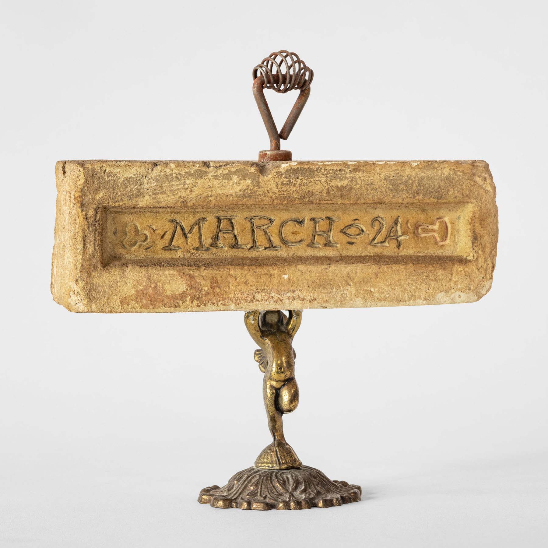 A decorative metal base that supports a cherub holding up a stone brick with “March 24” carved into it.