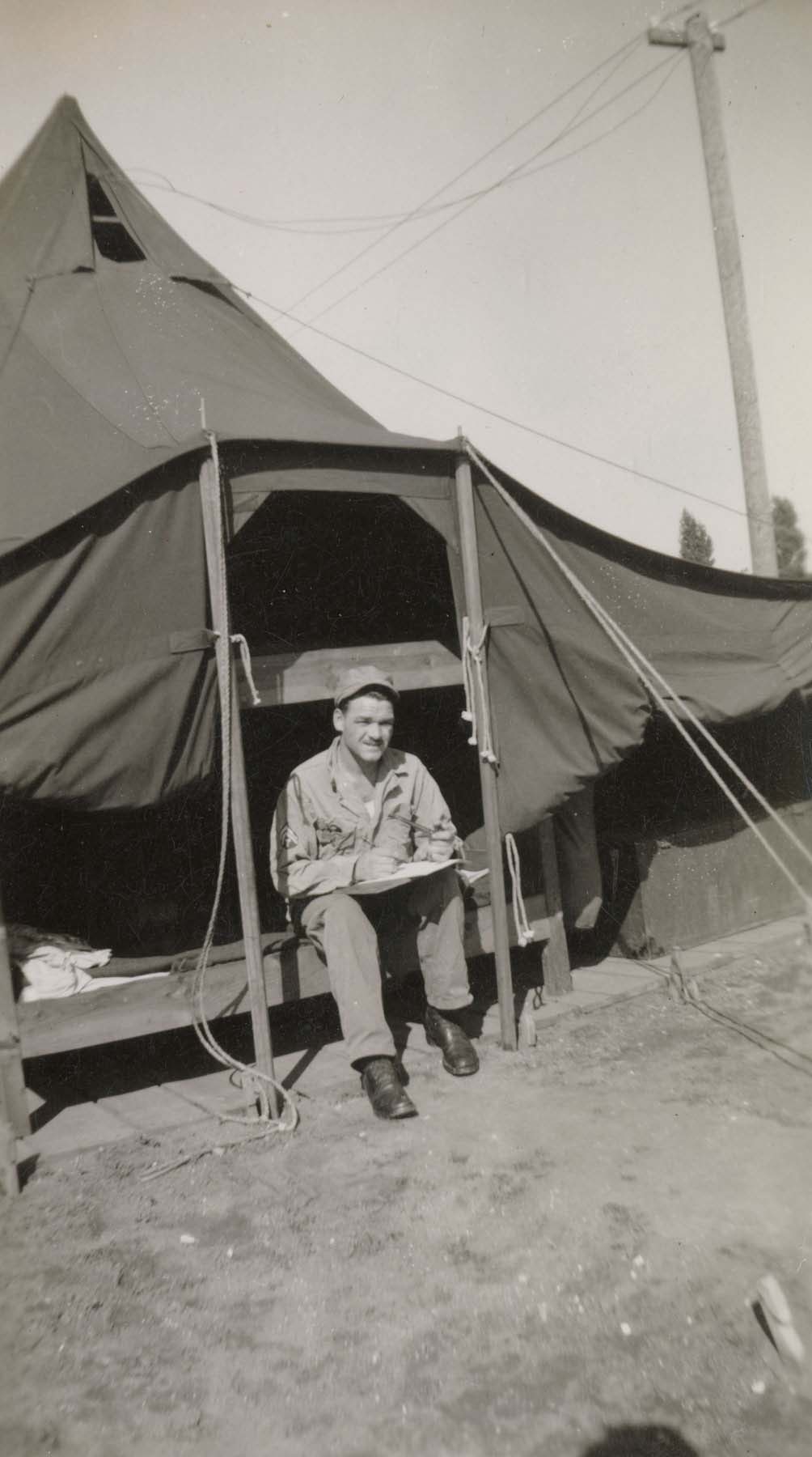 A black-and-white photograph of artist Charles T. Williams sitting and drawing in the opening of a military tent.