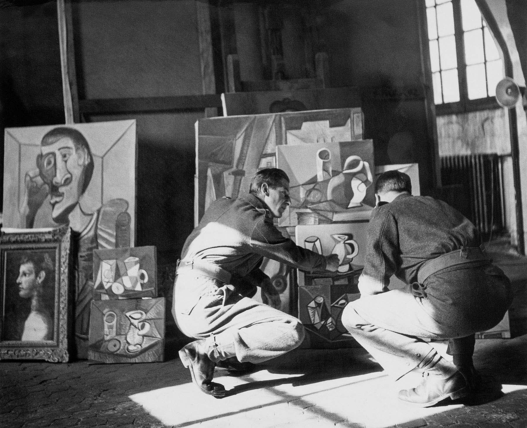 A black-and-white photograph of two men in military uniforms crouching in front of paintings in an artist's studio.