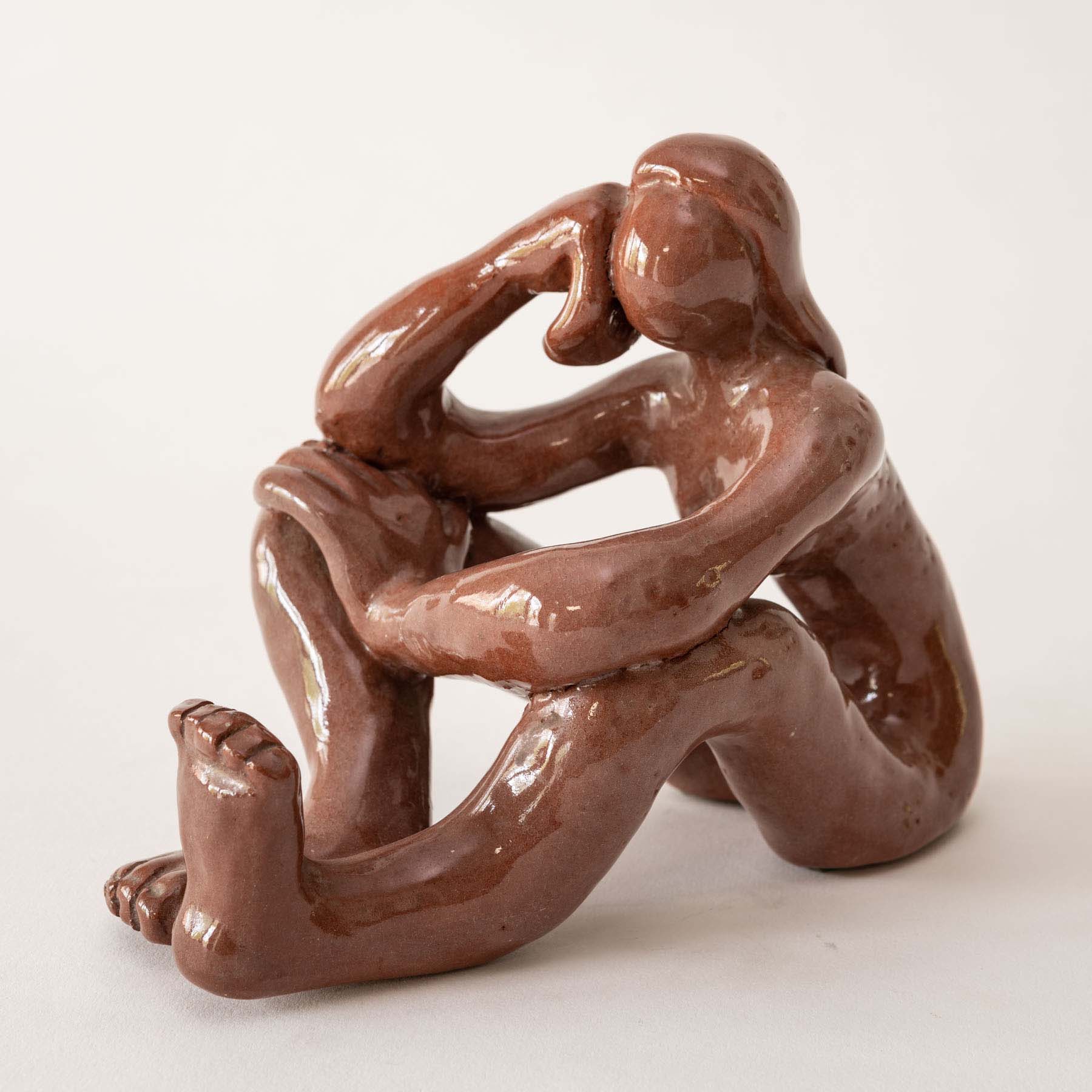 A brown, ceramic female seated figure rests her head on her hands and her elbows on bent knees.