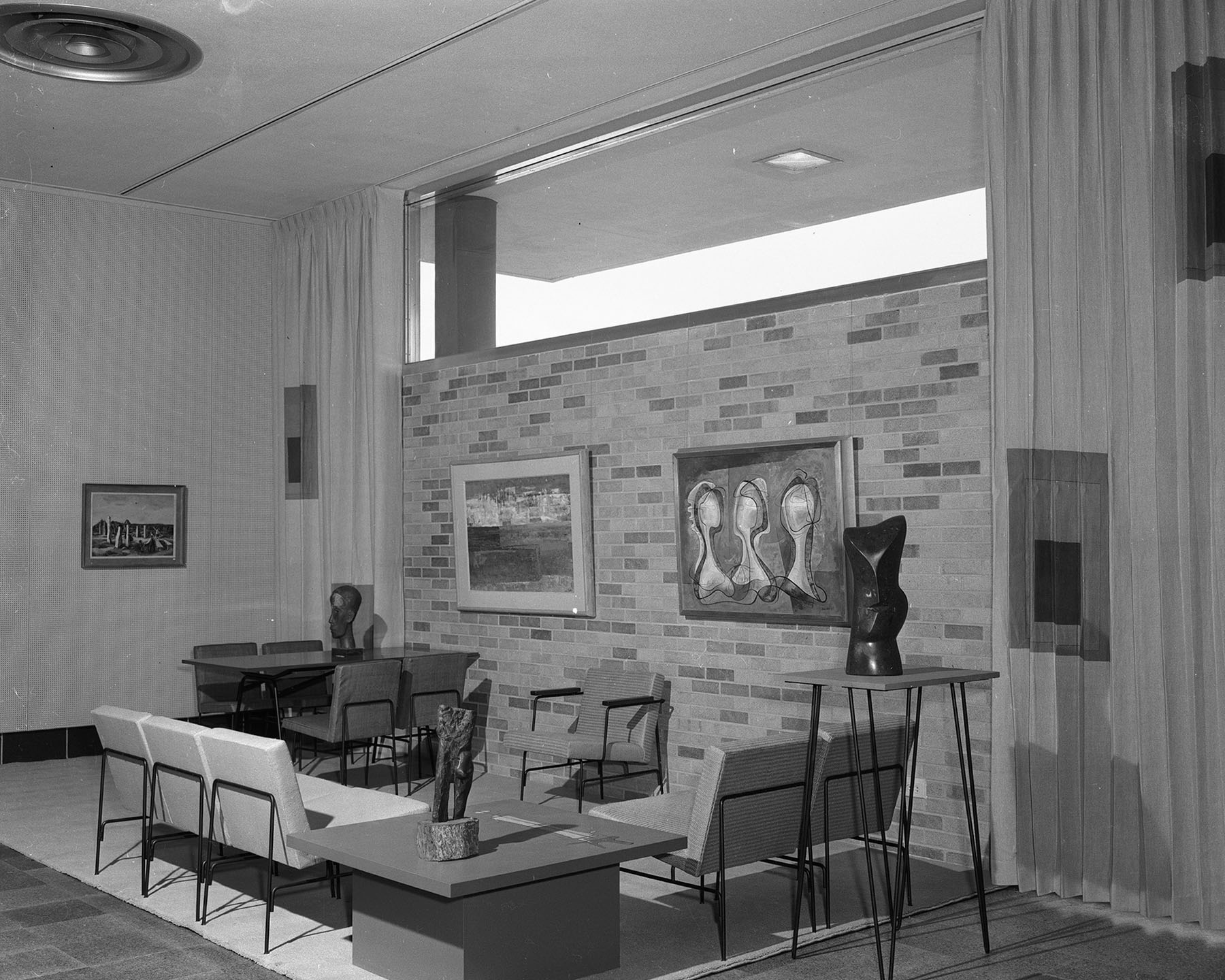 Black-and-white photo of a seating area with several sculptures displayed on tables and paintings on the wall.