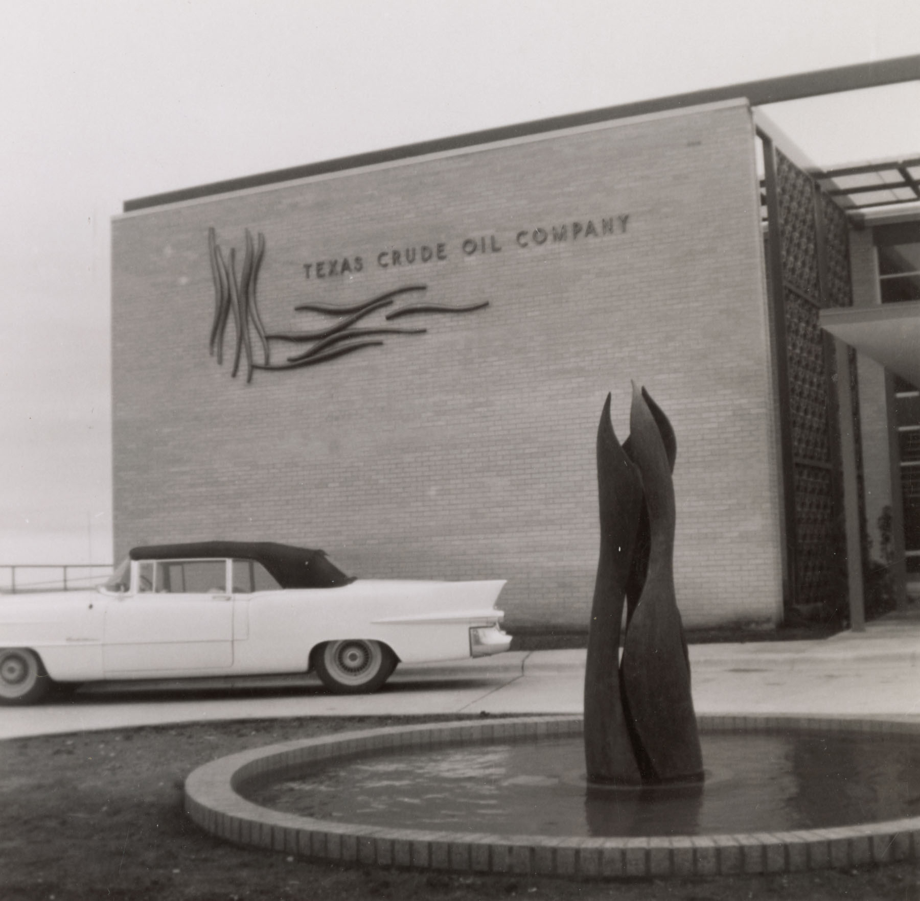 Black-and-white photo of the façade of the “Texas Crude Oil Company” and fountain with a tall sculpture in the center.