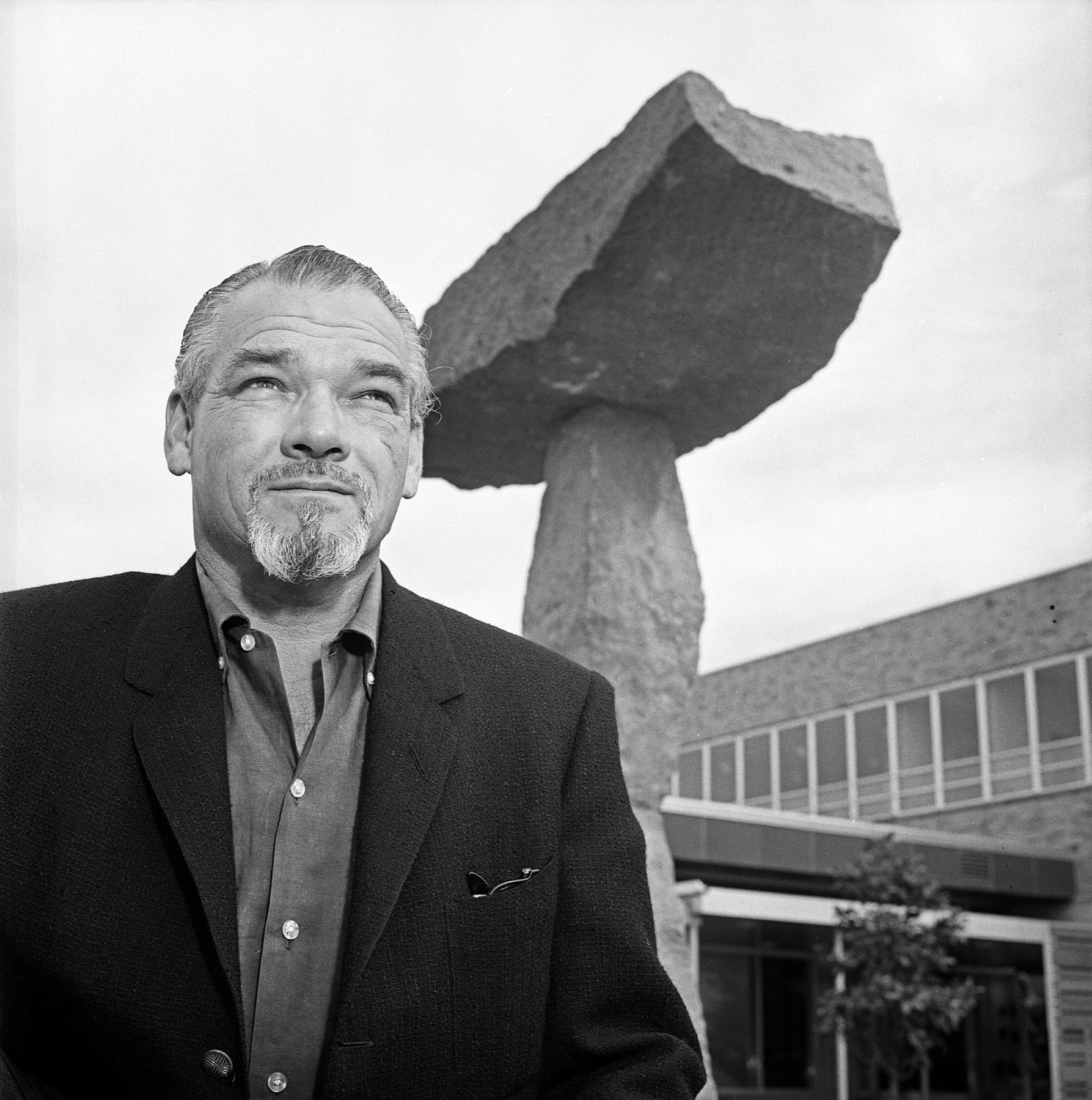 Black-and-white photo of a man with a goatee sitting under a tall stone sculpture.