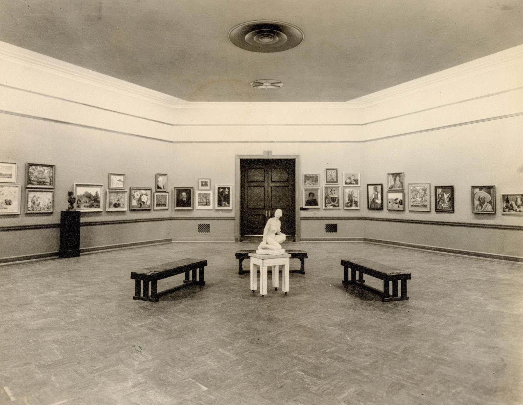 Black-and-white photo of a gallery with artwork on the walls and benches in the center arranged around a sculpture on a pedestal.