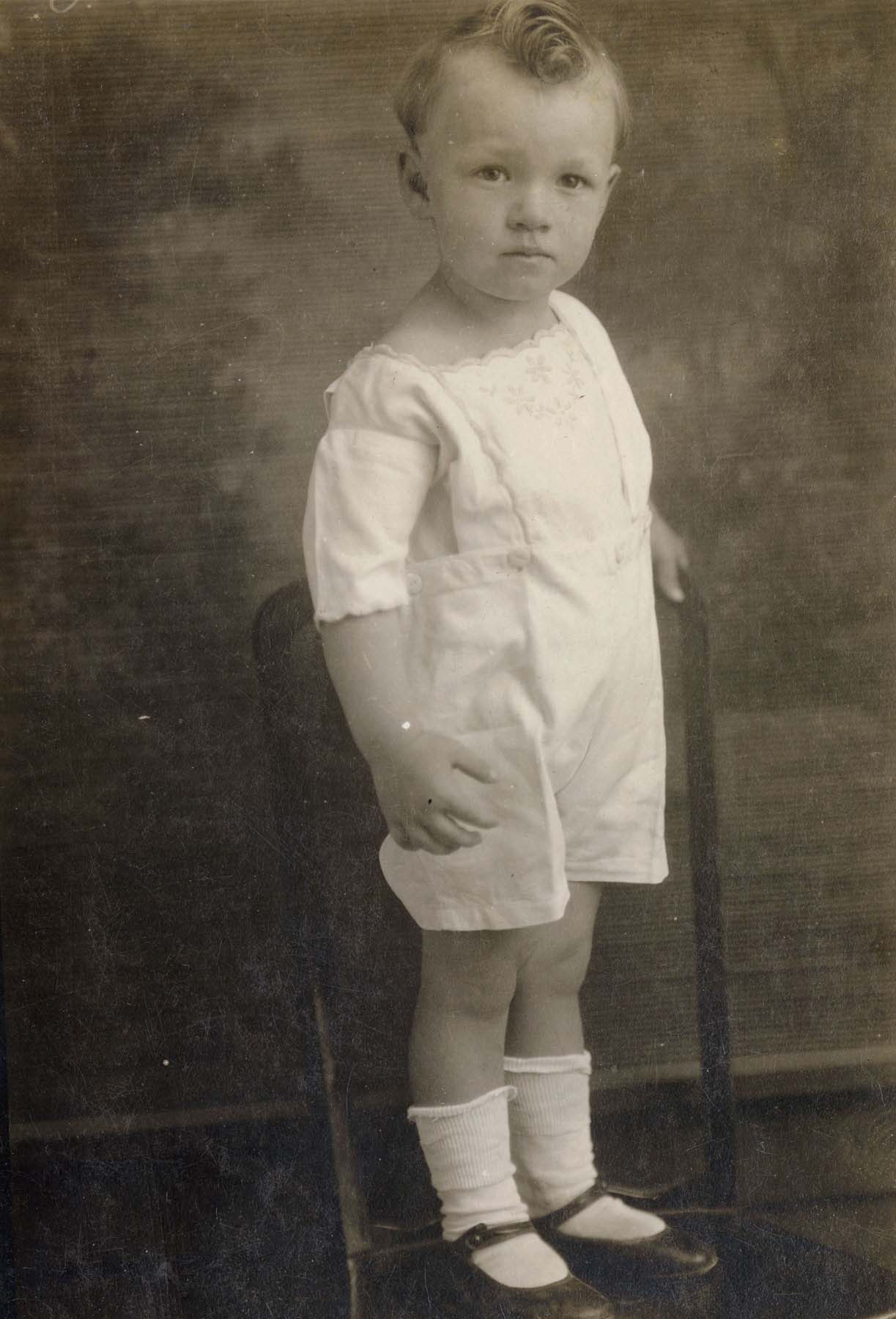 Formal black-and-white photo of a child standing on a chair.