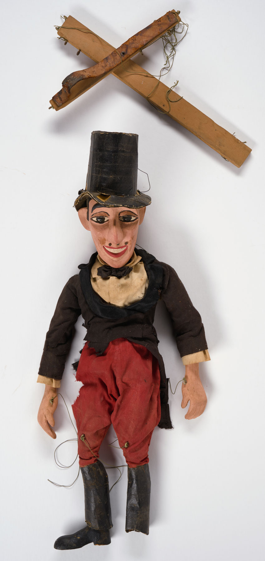 An aged marionette in a black top hat and jacket, red pants, and tall black boots, one of which is missing a foot.