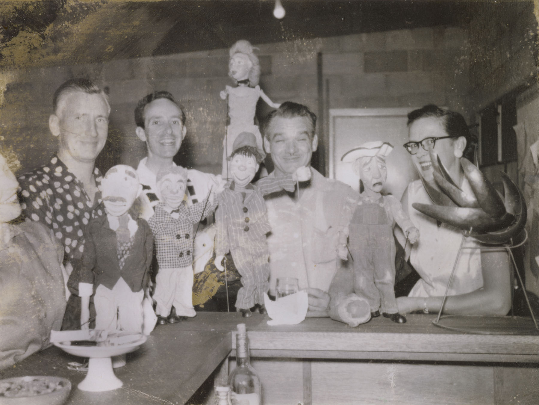 Faded photo of four people standing behind a counter on which are propped four marionettes and a sculpture.