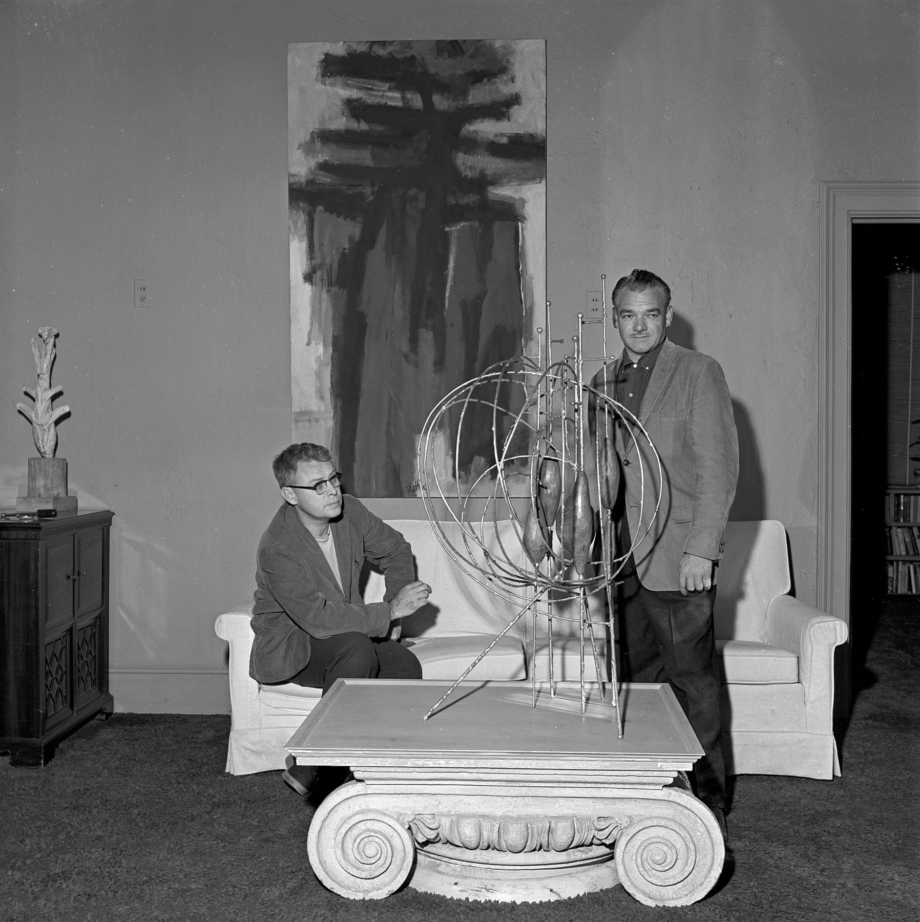 Black-and-white photo of a man seated on a sofa under a painting on the wall and another man standing by a coffee table on which a large sculpture sits.