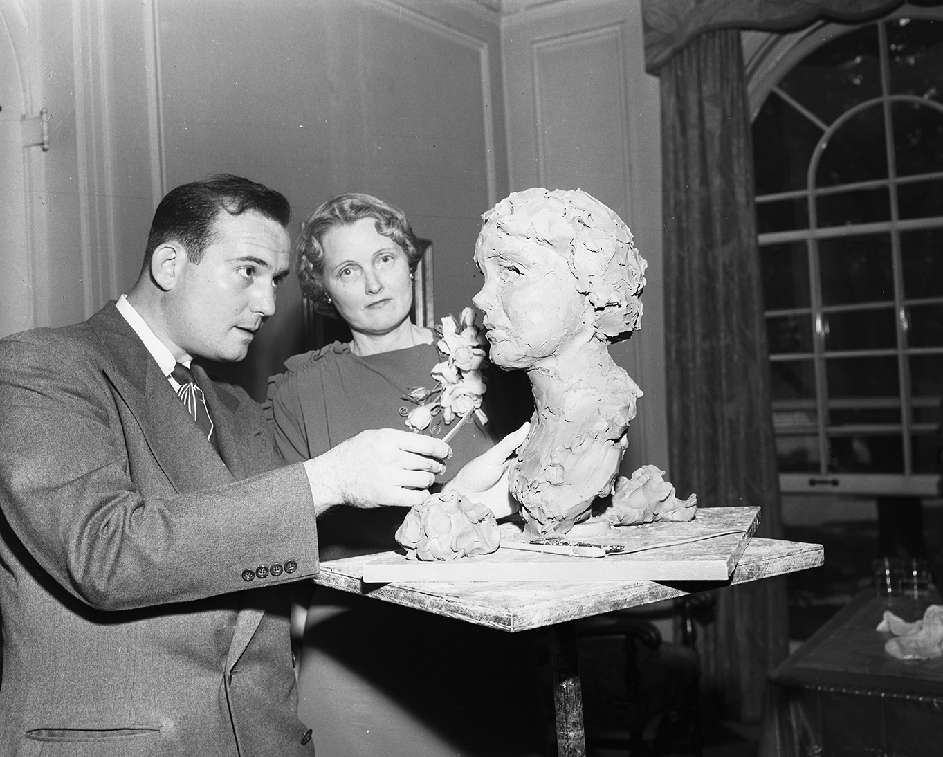 Black-and-white photo of a man sculpting a bust of the woman standing next to him.