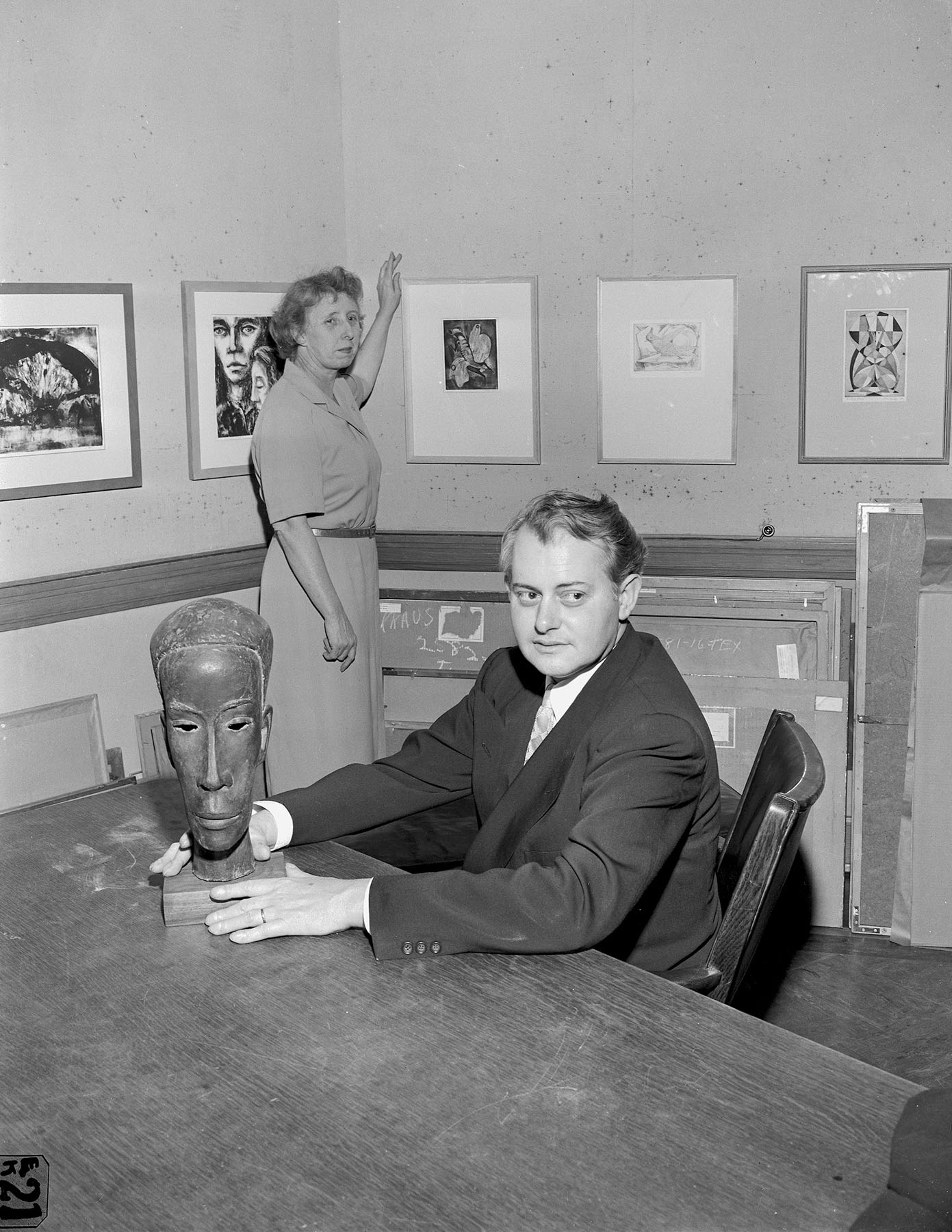 Black-and-white photo of a woman standing next to framed artwork on the wall and a man sitting at a table holding a sculpture.