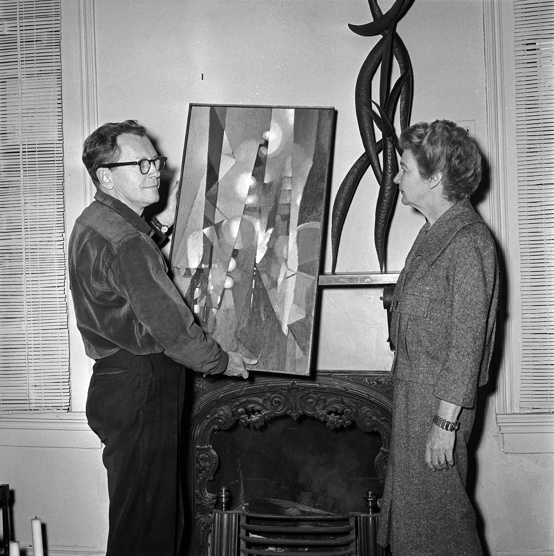 Black-and-white photo of a man holding a painting and a woman standing next to a sculpture on a mantel.