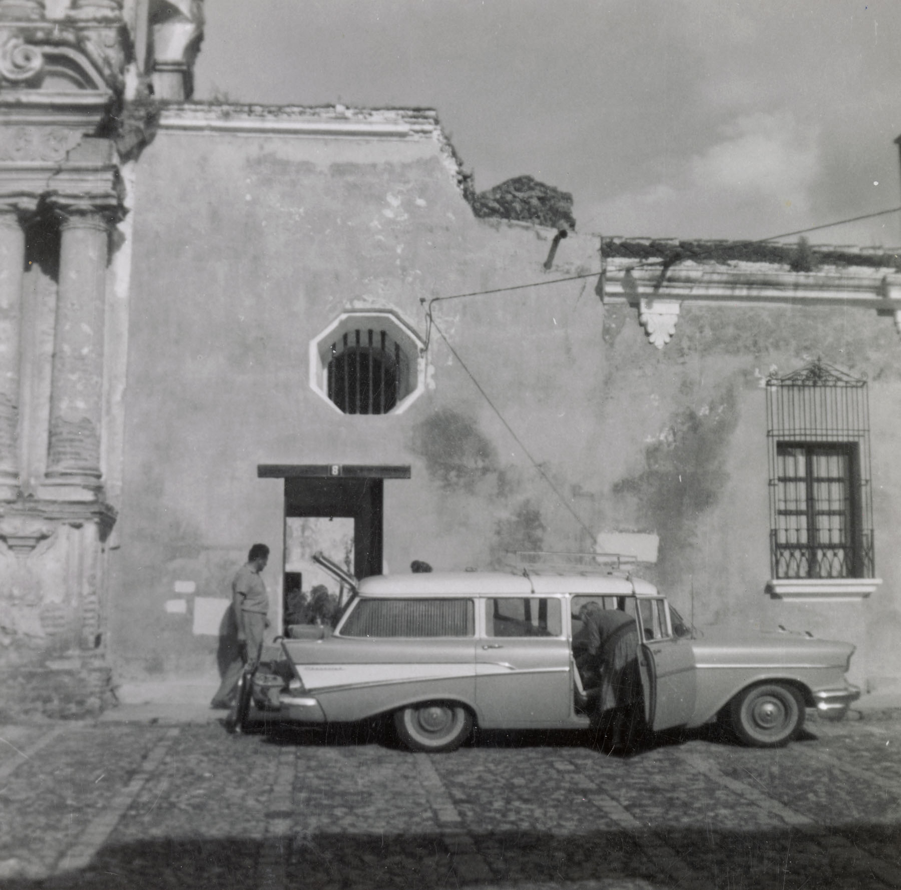 Black-and-white photo of two people standing near a 1950‘s style station wagon parked in front of a building.