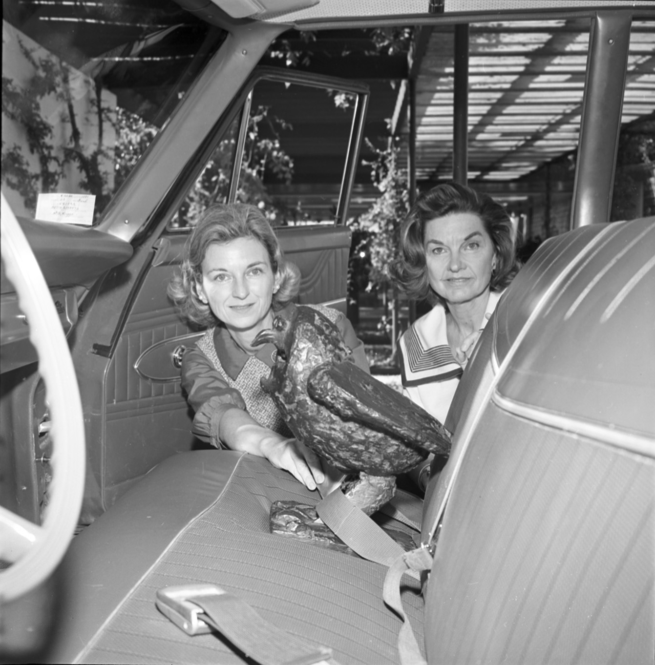 Black-and-white photo of two women kneeling next to an open car door placing a seatbelt on a sculpture of a bird.