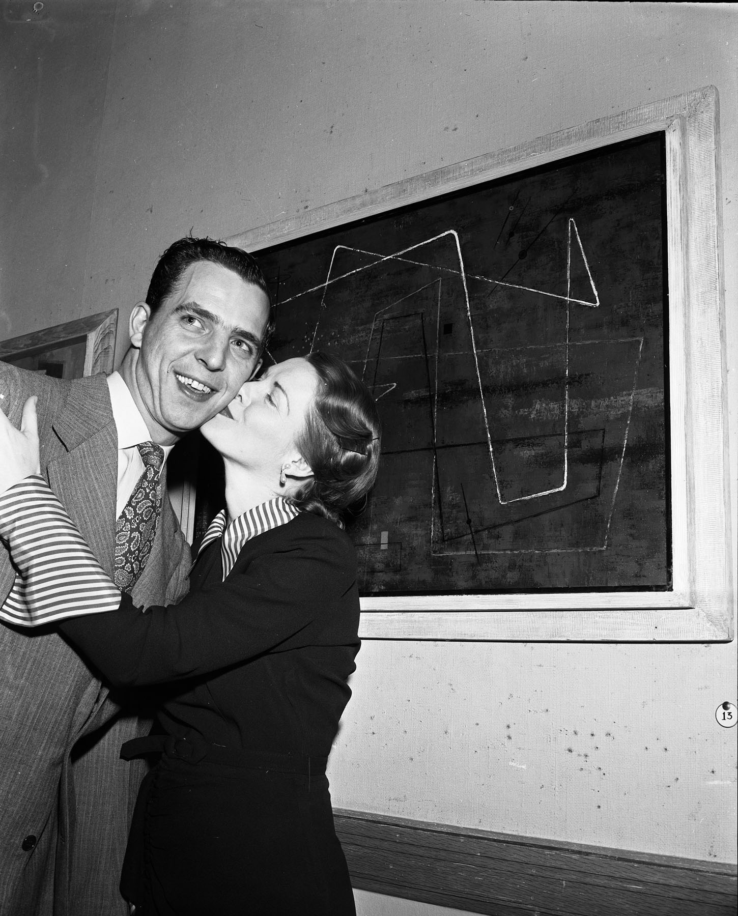 Black-and-white photo of a woman kissing a man on the cheek as they stand in front of a painting.