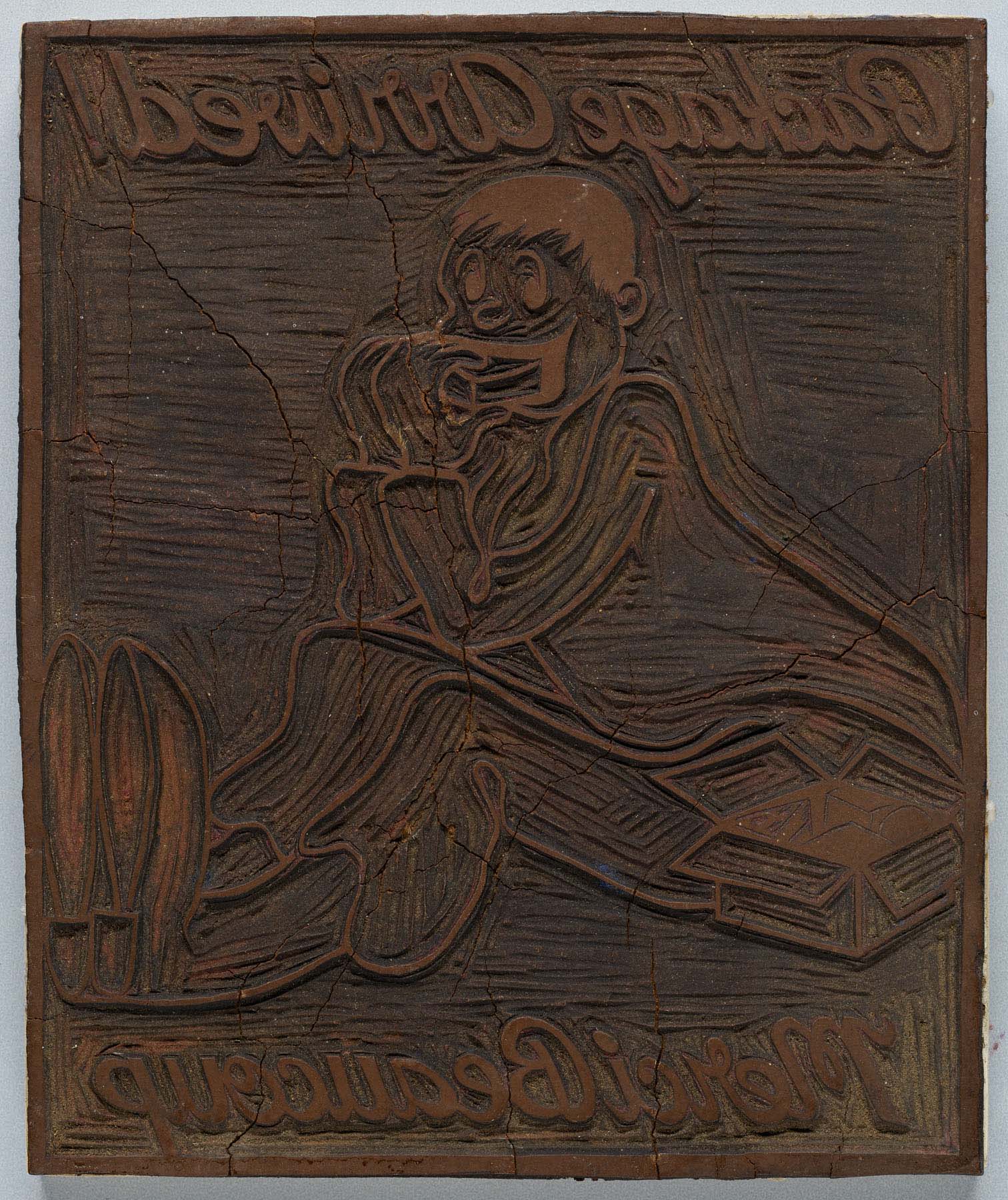 A carved wood block showing an image in reverse of a seated man next to an open package with the words “Package Arrived! Merci Beaucoup.”