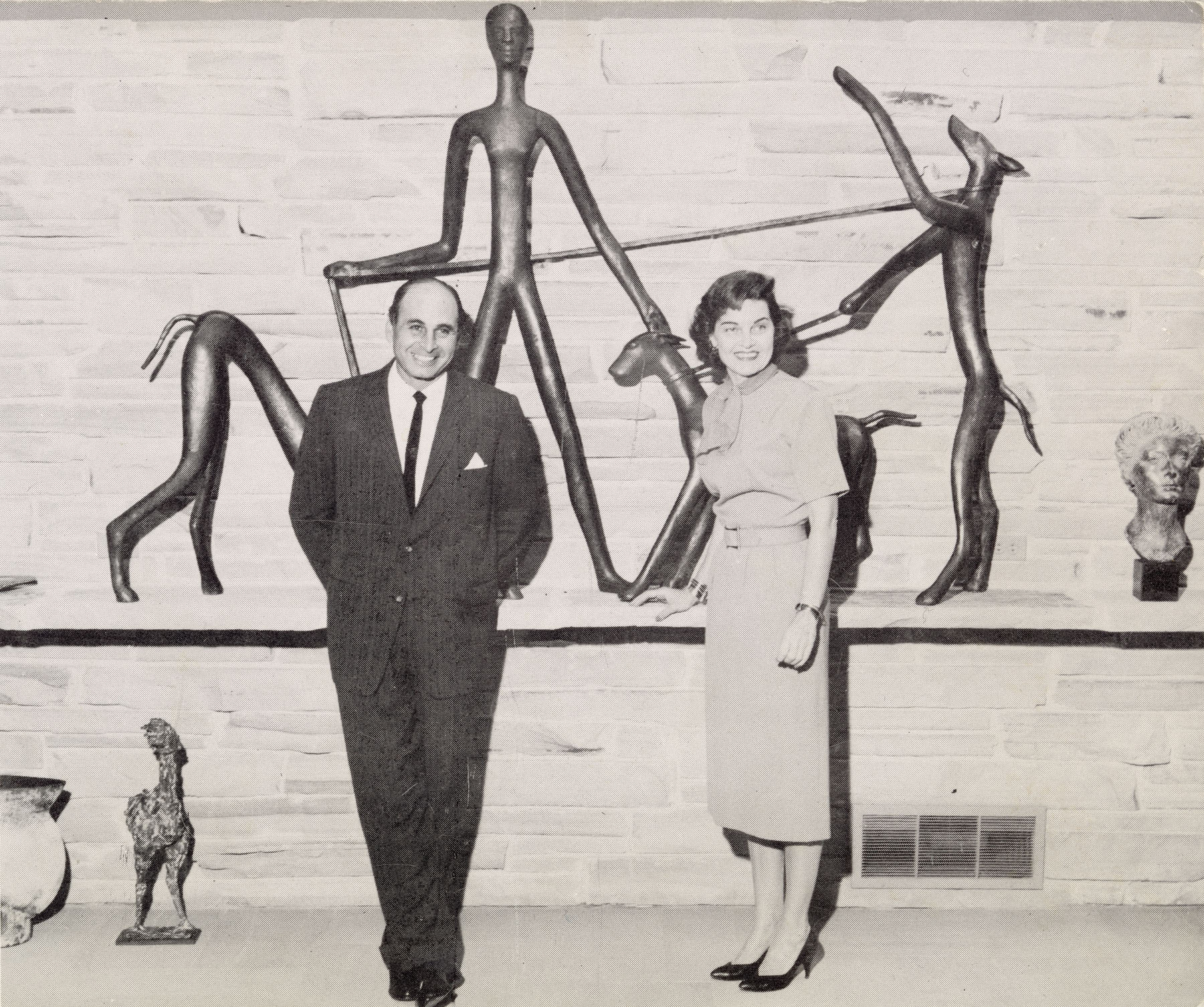 Black-and-white photo of a man and a woman standing in front of a stone wall on which several sculptures are displayed.