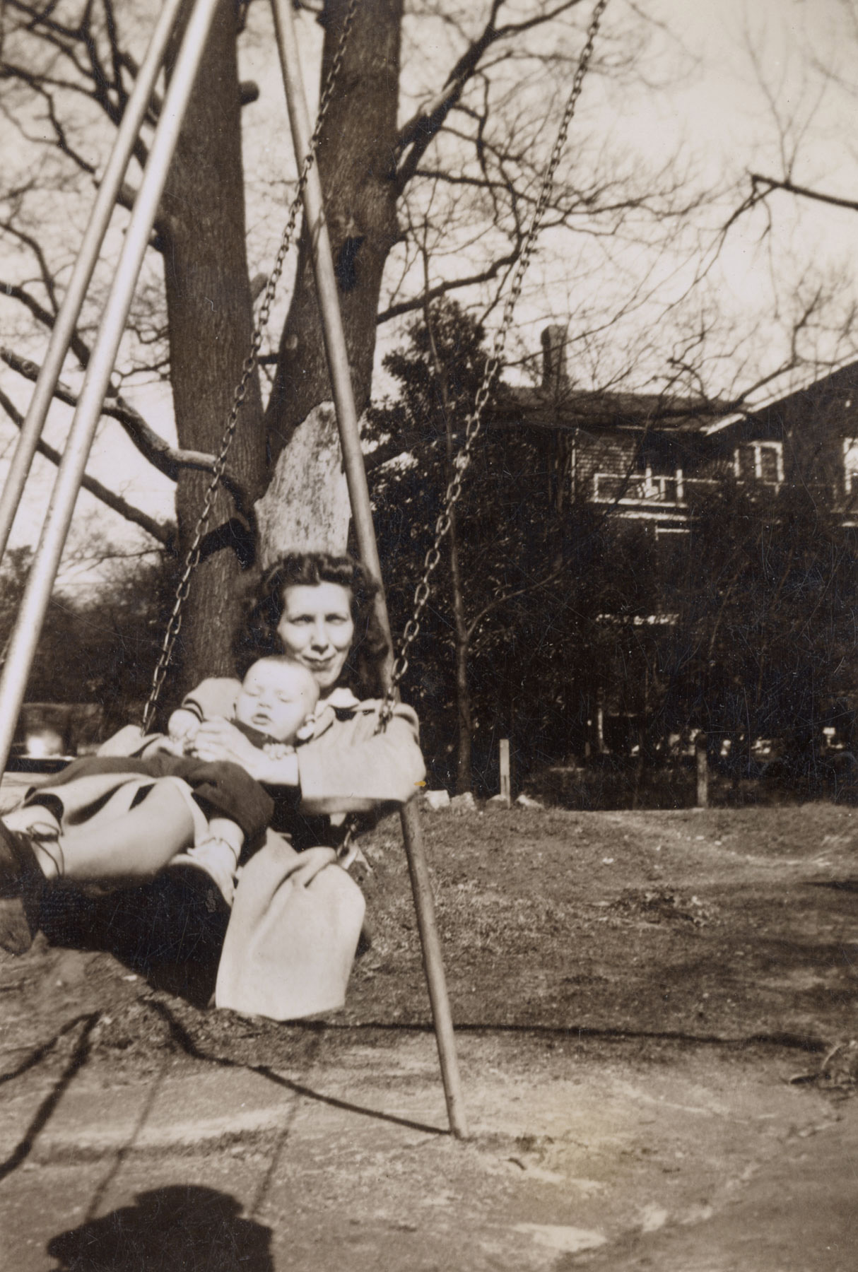 Black-and-white photo of a woman holding a baby in her lap as they swing on a swingset.